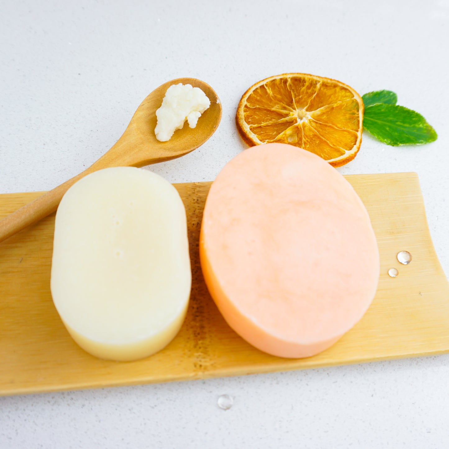 A large sweet orange shampoo bar and a conditioner bar on a wooden bamboo dish. A wooden spoon with shea butter in the background